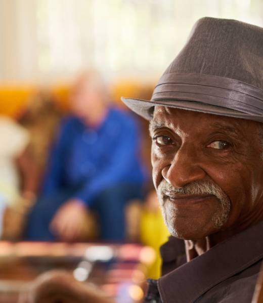Portrait of elderly black man looking at camera in retirement home, with group of friends in background. Patients relaxing in hospice for seniors.
